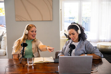 Diverse female co-hosts recording podcast in living room at table with laptop microphones