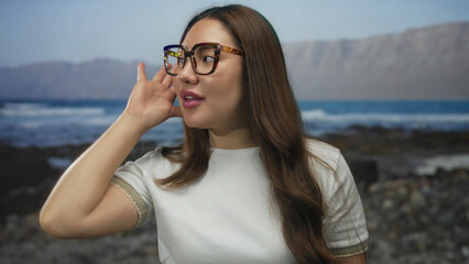 Woman adjusts tortoiseshell eyeglasses with hand to ear while gazing toward sunlit ocean waves on beach; listening inquiry attention curiosity.