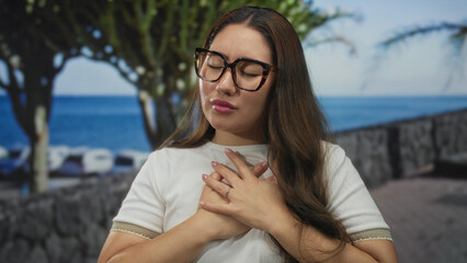 Young hispanic woman wearing glasses and white t shirt clutching chest on sunny seaside promenade by a stone walkway; pain.