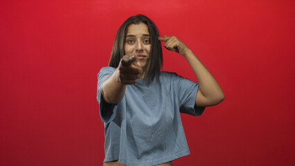 Young hispanic woman pointing finger to camera in red studio midshot with relaxed pose and casual tee; playful defiance.