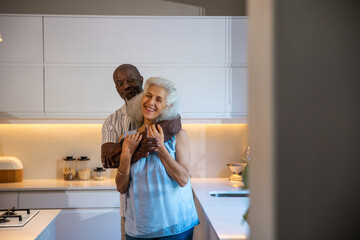 Diverse senior couple hugging in modern kitchen featuring glass jars bread box and gas cooktop
