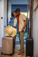 African American couple embracing in entryway by front door, holding sunhat near suitcases