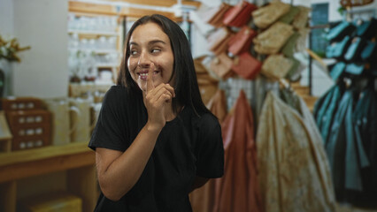 Young hispanic woman with finger to lips gesture in a clothing shop with racks and purses visible; quiet secrecy.