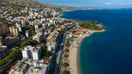 Fototapeta premium Aerial view of Messina seafront, Sicily, Italy. Panorama of a coastal city nestled between green hills and the blue sea. A long sandy beach borders the urban landscape under a clear Mediterranean sky.