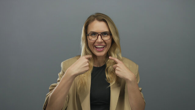 Woman smiling and gesturing at herself in a beige blazer against a plain grey background.