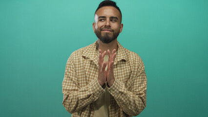 Young hispanic man with hands pressed together and a slight smile wearing a checked shirt in a turquoise studio; calm gratitude.