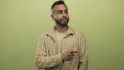 Fototapeta premium Man pointing finger in studio against olive green backdrop wearing checked shirt and rings on hand; thoughtful curiosity.