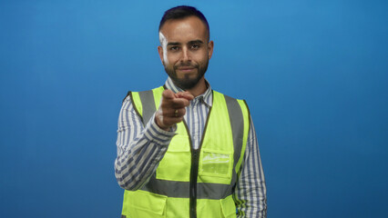 Fototapeta premium Man pointing finger in studio wearing reflective safety vest as engineer against blue backdrop; confidence recruitment.