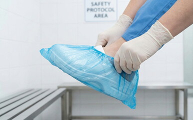 Person wearing medical gloves puts on a disposable blue shoe cover, adhering to hygiene and safety protocols in a sterile environment.