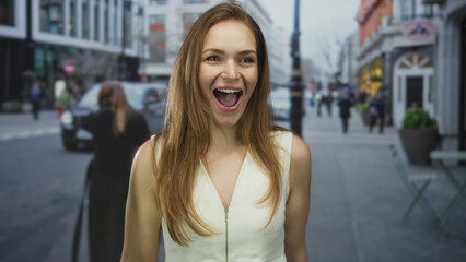 Fototapeta premium Young woman with open mouth laughing and showing teeth amid blurred pedestrians on a sunny urban street scene in daytime; joy.