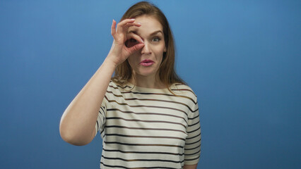 Woman wearing striped shirt frames one eye with binocular hand gesture in blue studio; curiosity...
