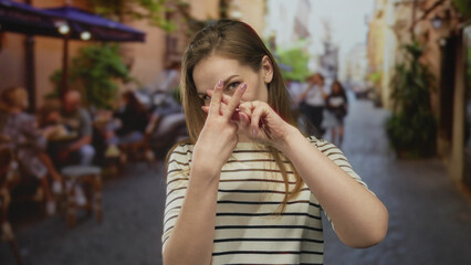 Woman crossing index fingers in front of her on a busy street terrace by cafe tables under umbrellas with a playful smile; rejection.
