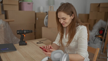 Woman taps smartphone on wooden table in building among moving boxes and power drill; calm preparation.