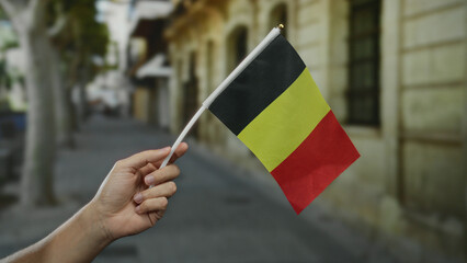 Man's hand holding a small belgian flag in an outdoor street setting in a city.