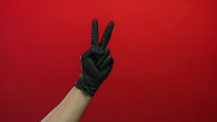 Hand of a man wearing black gloves showing a peace sign against a vibrant red background, highlighting a sense of isolation and focus over the striking wall.