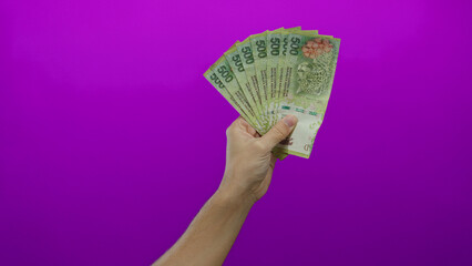 Man holding argentine pesos in hand with a vibrant purple background, showcasing currency in an isolated setting for financial context.