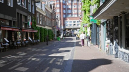 Obraz premium Blurred people walking down a sunlit street in amsterdam's urban environment with defocused buildings and cafes creating a bokeh effect.
