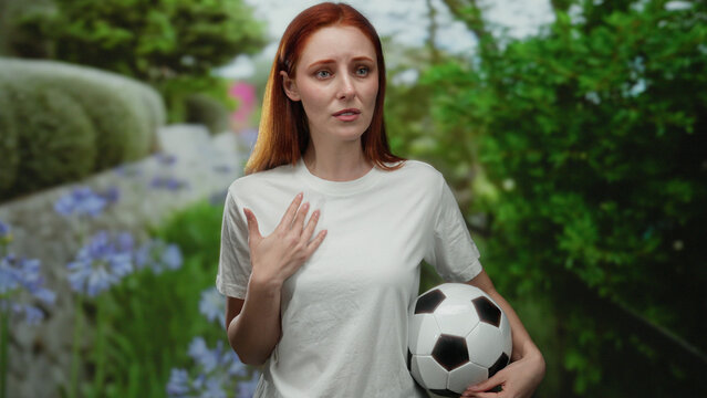 Woman with red hair in a park holding a soccer ball while wearing a white shirt surrounded by greenery and vibrant flowers, expressing surprise or confusion.
