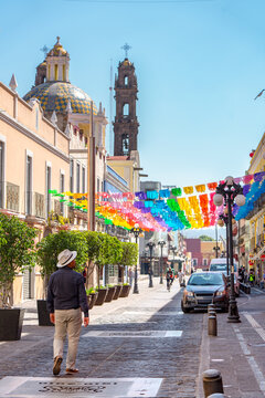 A man wearing a white hat walks down a sunlit cobblestone street in Puebla, Mexico, which is decorated with vibrant banners and flanked by historic buildings featuring a prominent tiled dome and bell 