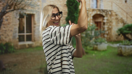 Woman with bare arm raises arm and smiles wearing striped shirt and oversized glasses in front of old stone building; carefree joy.