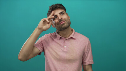 Young man points finger to temple in studio while gazing at camera with slight head tilt; thoughtful.