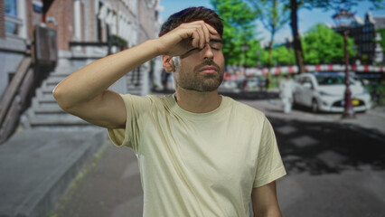 Fototapeta premium Man wipes sweaty forehead on sunlit city street with brick building steps and parked car behind; fatigue heat.