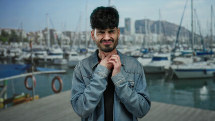 Young man with throat pain stands outdoors near a port with boats and water in the background,...