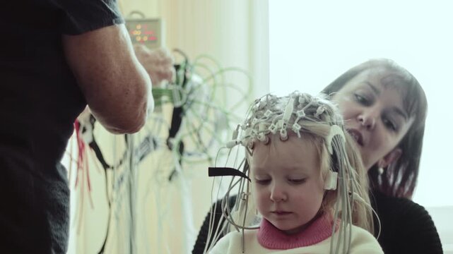 Doctor meticulously applies electrodes to head of 4 years old child patient, beginning electroencephalography examination. Brain electrical activity for medical diagnosis. Mother parent