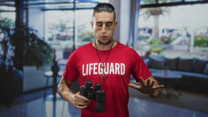 Lifeguard man holding binoculars with raised right hand and whistle around neck, focused gaze, hands visible in building lobby with seating and plants; duty vigilance.