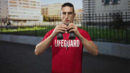 Man holding binoculars to eyes with whistle visible, wearing red lifeguard shirt and standing by a...
