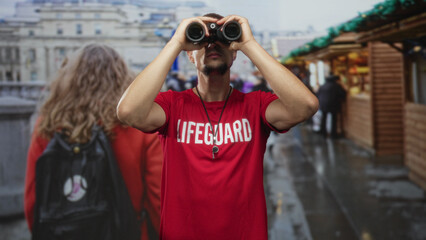 Lifeguard man in red shirt holds binoculars to eyes and peers along a wet street market; vigilance...