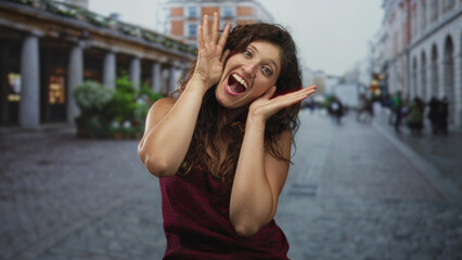 Fototapeta premium Young hispanic woman laughing with hands to face and covering eyes on a crowded cobbled street beside a colonnade of shops; joy playful surprise.