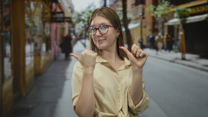 Young blonde woman with thumbs pointing to chest wearing glasses and a beige shirt on a busy city street; confidence pride positivity empowerment.