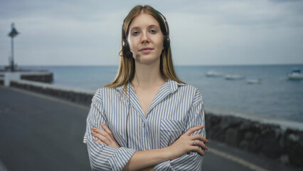 Woman wearing a headset microphone crossing her arms on a street beside the sea under an overcast sky; confidence.