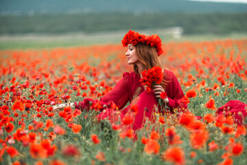 Woman poppies field sitting in long red dress wearing floral crown on a summer day