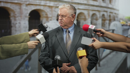 Senior man in suit with clasped hands and closed eyes facing multiple handheld microphones from reporters in front of an ancient colosseum on a street  concern. © Krakenimages.com