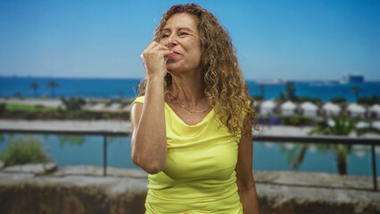 Woman smiling and raising hand in a pinched fingers gesture, wearing yellow sleeveless top and necklace on a studio seaside set; joyful.