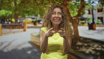 Middle aged hispanic woman in yellow shirt pointing index fingers at camera on street plaza lined with trees and benches; playful surprise.