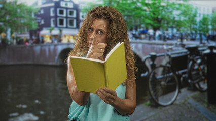 Middle-aged woman holds yellow book and places two fingers to face while reading beside a canal and bicycles on a street; contemplation.