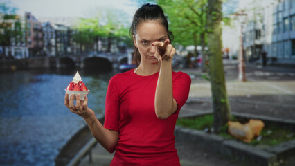 Woman holding strawberry cream cup points finger to cup on a street by a canal bridge walkway;...