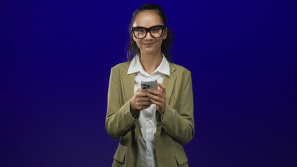 Woman in olive blazer holding smartphone with both hands, looking down and typing on the screen while standing in a studio with deep blue backdrop; concentration.
