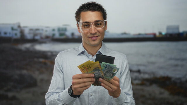 Young man with glasses counting australian dollars from wallet by the seaside, smiling and wearing a watch, embodying a casual beach setting.