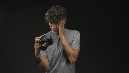 Teen boy holding dslr camera inspecting images, hand on cheek for toothache in studio; frustration uncertainty.