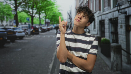 Teenage boy pointing finger upward, arms crossed, standing on a city street by parked cars and stone building; quiet curiosity.