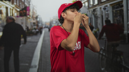 Lifeguard man in red shirt and cap blowing a whistle and raising both arms on a busy street,...