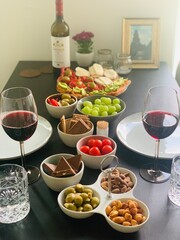 Tapas at Home with Wine and Shared Snacks on Dining Table; Cozy Home Tapas Table with Wine and Shared Appetizers; olives, grapes, cheese, crackers, chocolate, nuts, tomatoes, and open sandwiches