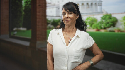 Woman senior with hand on hip wearing white blouse and necklace, looking sideways beside stone colonnade in building garden; confidence.