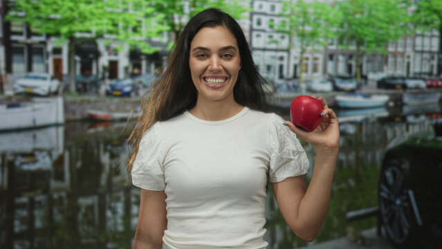 Woman cringes while holding red apple by amsterdam canal street with historic buildings visible; distaste.
