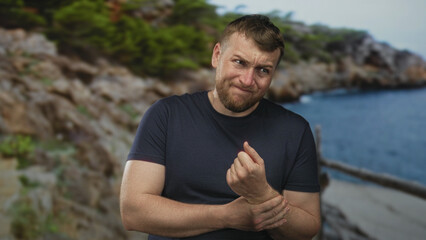Man clutching wrist and grimacing while holding forearm on a coastal forest path by rocky cliffs and sea, wearing a dark tshirt; pain recovery.