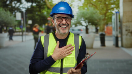 Man smiling in urban street wearing hardhat and safety vest while holding clipboard with colleagues...